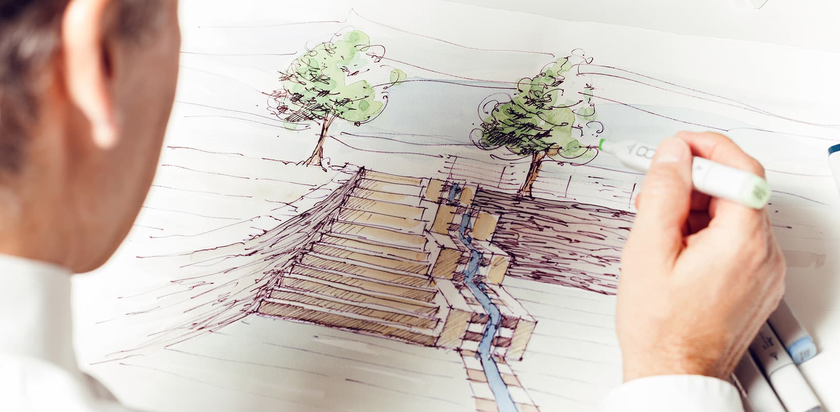 Eine Person skizziert mit einem Marker Architekturpläne mit Treppen, Bäumen und einem Wasserspiel.